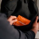An orange Coffeeangel brew bag being slipped into the front pocket of a black backpack.