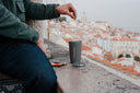 Someone sitting on a stone wall above a city, lowering a brew bag into a grey Coffeeangel tumbler.