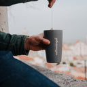 A person holds a dark gray insulated travel mug with the brand name 'coffeeangel' printed vertically in white. A coffee brew bag is being steeped into the mug, with a blurred coastal cityscape in the background.