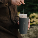 Lowering a Coffeeangel brew bag into a grey insulated tumbler held in one hand outdoors.