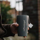 A grey Coffeeangel tumbler held out at arm's length with steam rising and a brew bag tag hanging over the rim.