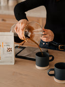 Two contemporary stoneware mugs on a kitchen counter as coffee is poured from a Chemex